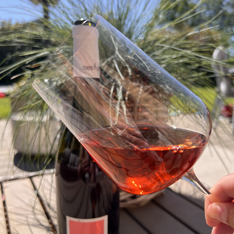 A single elegant wine glass filled with Tenuta Demaio Nero di Troia Rosato, showcasing its delicate cherry-pink color on a rustic wooden outdoor table with a sun-drenched vineyard background.