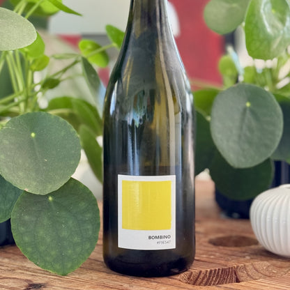 Bottle of Bombino wine with a yellow label on a wooden surface surrounded by green plants.