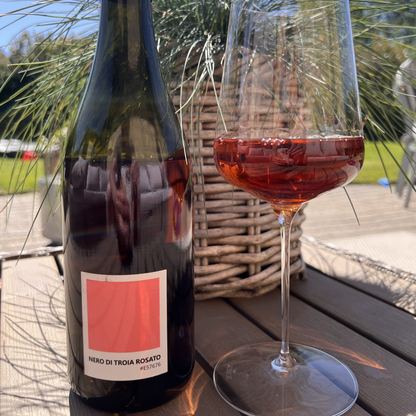 A close-up lifestyle shot of a hand holding a bottle of Tenuta Demaio Rosato next to a poured glass of the cherry-pink wine on an outdoor wooden table, emphasizing a relaxed dining atmosphere.