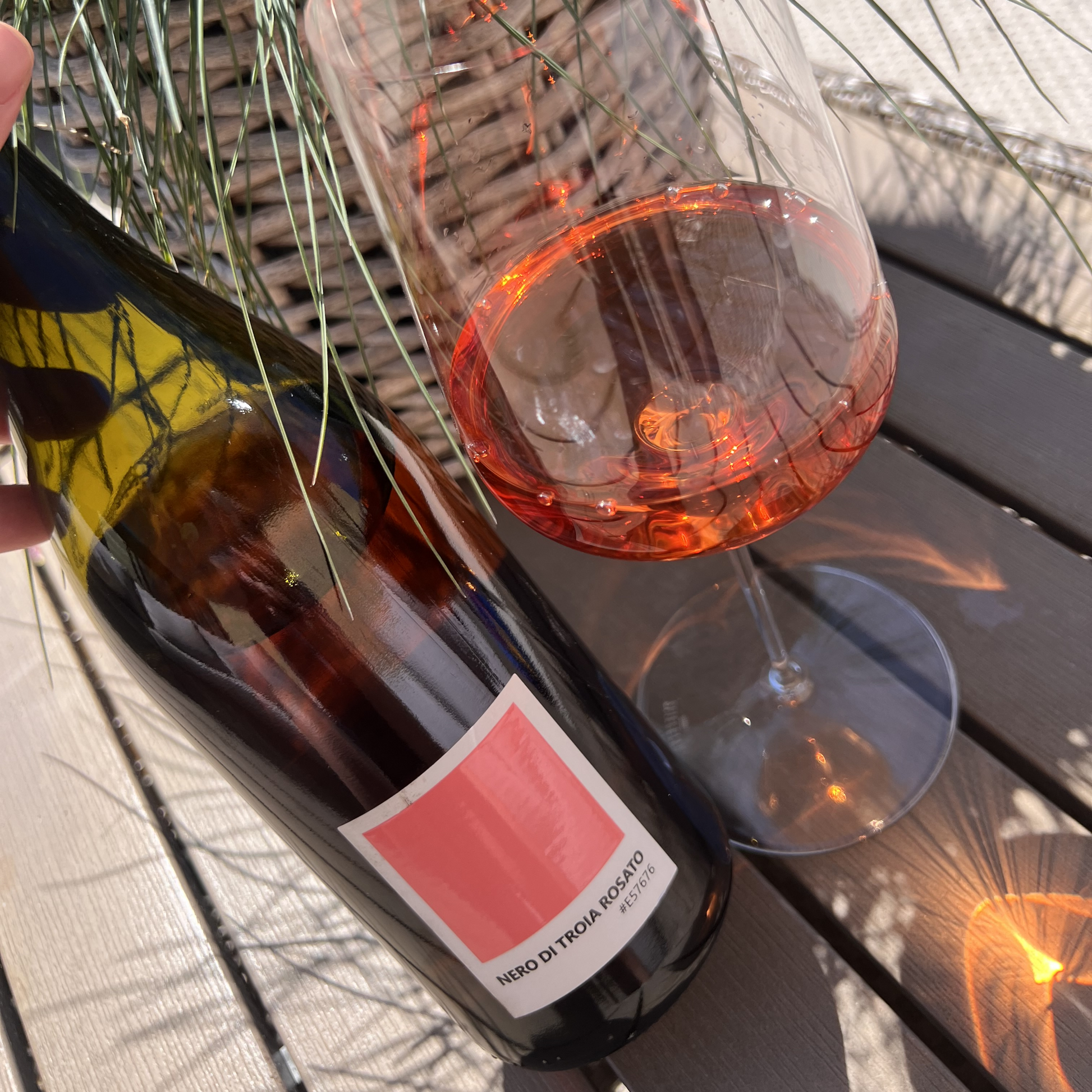 A bottle of organic Tenuta Demaio Nero di Troia Rosato standing next to a filled wine glass on a weathered wooden table, capturing the unfiltered rosé in bright, natural outdoor light.