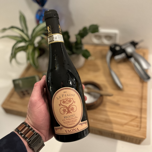 A close-up shot of a hand holding a bottle of Vaona Recioto della Valpolicella Classico DOCG, set against a softly blurred rustic wooden cutting board in the background.