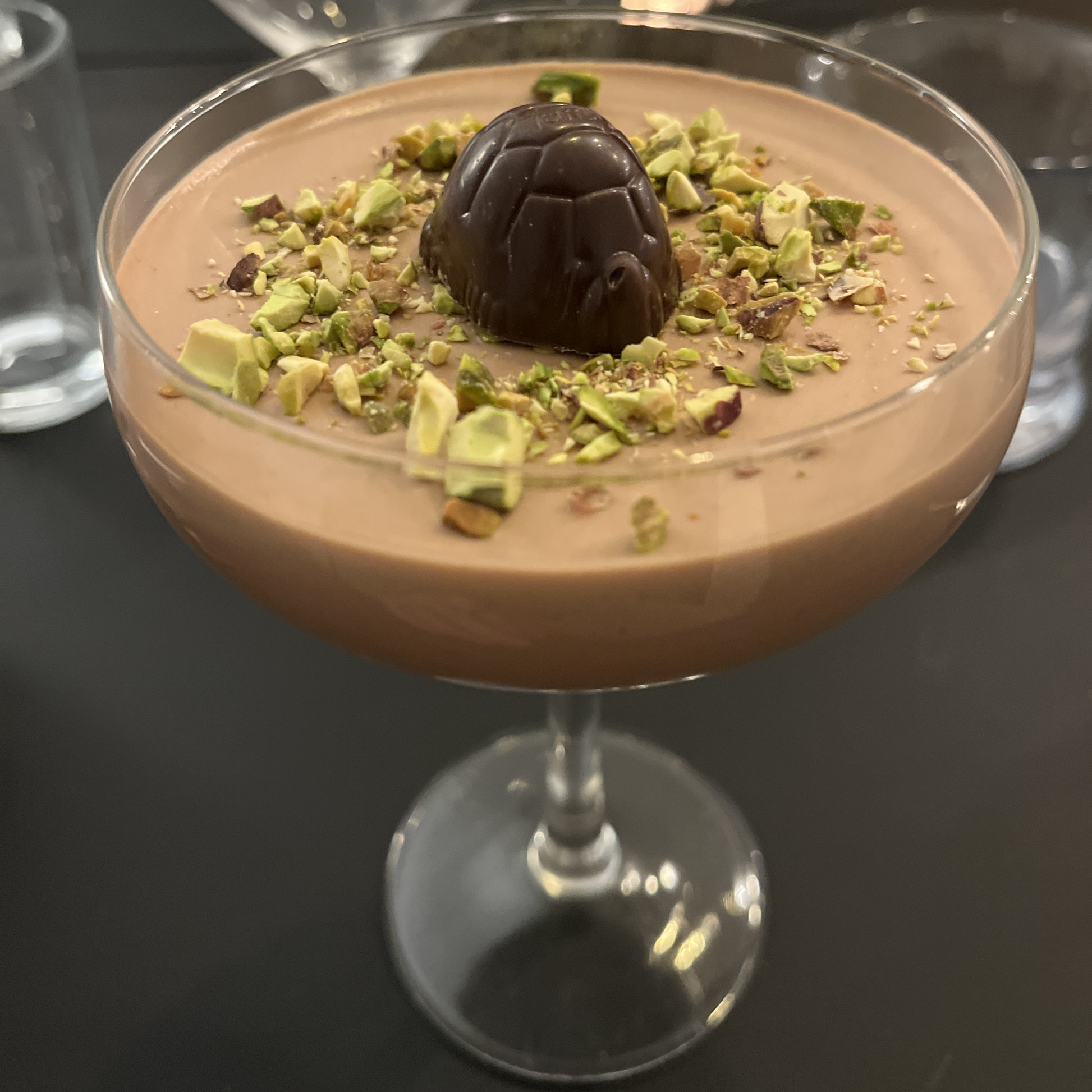 A detailed close-up of a chocolate mousse dessert served in a fancy glass, beautifully topped with green pistachio crumble and a classic Danish Skildpadde chocolate