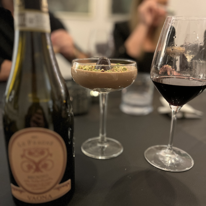 An elegant tabletop display featuring the chocolate mousse dessert and a filled glass of dark Recioto red wine side-by-side in sharp focus, with the Vaona Recioto bottle standing slightly blurred in the background.