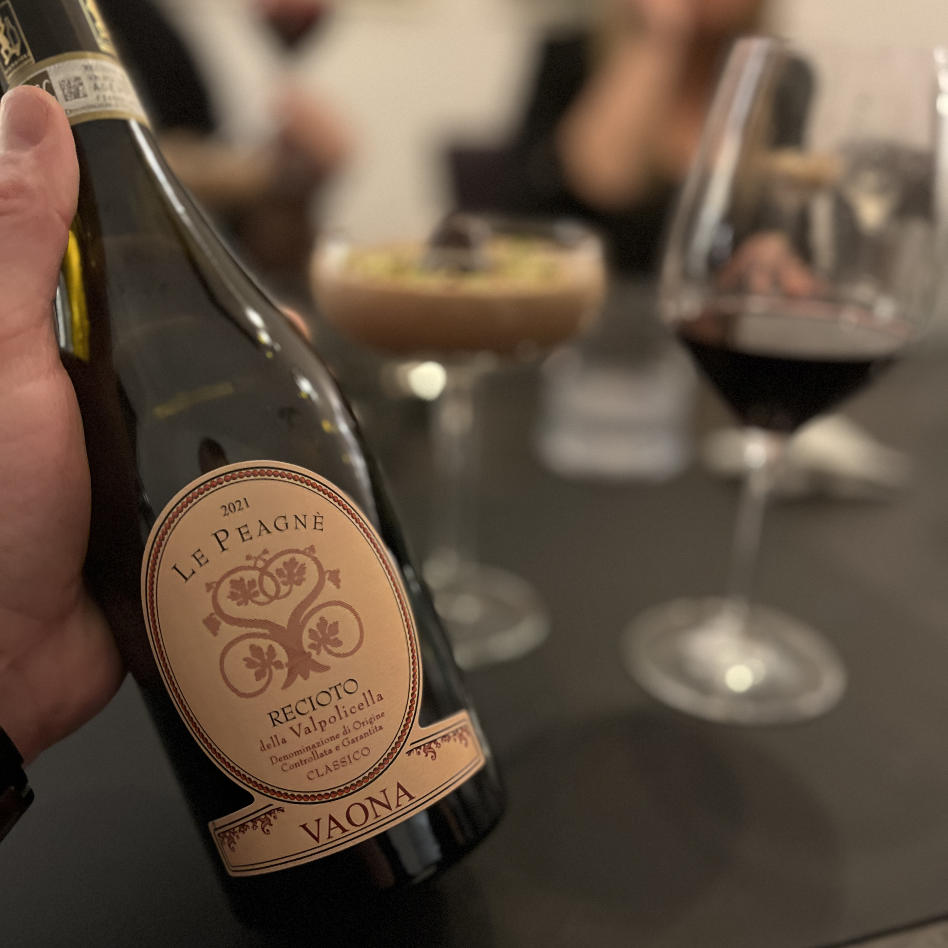 A product-focused shot highlighting the Vaona Recioto della Valpolicella bottle in sharp focus, while the chocolate mousse dessert and poured wine glass are artfully blurred next to it.