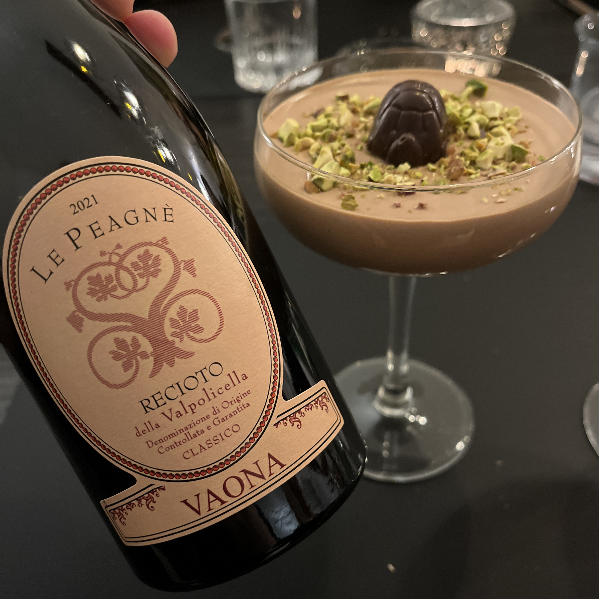 A lifestyle image showing a hand holding the bottle of Vaona Recioto right next to the chocolate mousse dessert topped with a Skildpadde chocolate, capturing a perfect dessert pairing moment without the wine glass.