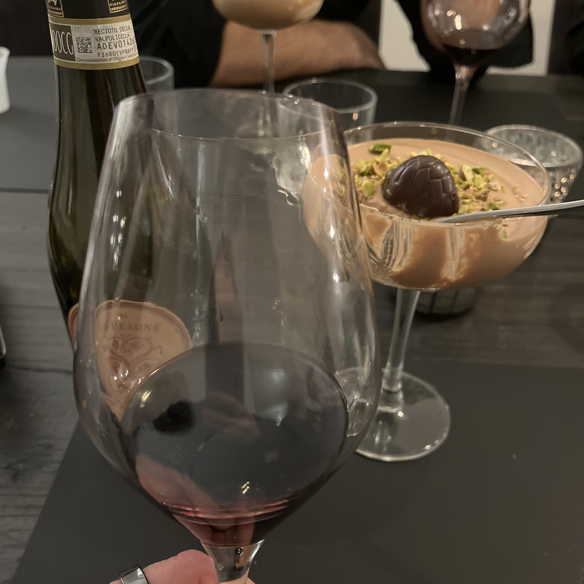 A front-focus shot highlighting a glass filled with deep red Recioto dessert wine to showcase its rich color, with the Vaona bottle and the chocolate mousse dessert positioned softly in the background.