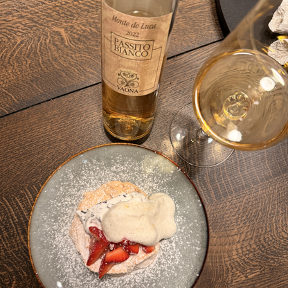 A lifestyle display featuring a bottle of Vaona Passito Bianco sweet wine and a filled dessert wine glass standing elegantly on a table next to a fresh strawberry dessert.