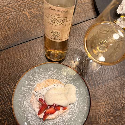 A lifestyle display featuring a bottle of Vaona Passito Bianco sweet wine and a filled dessert wine glass standing elegantly on a table next to a fresh strawberry dessert.