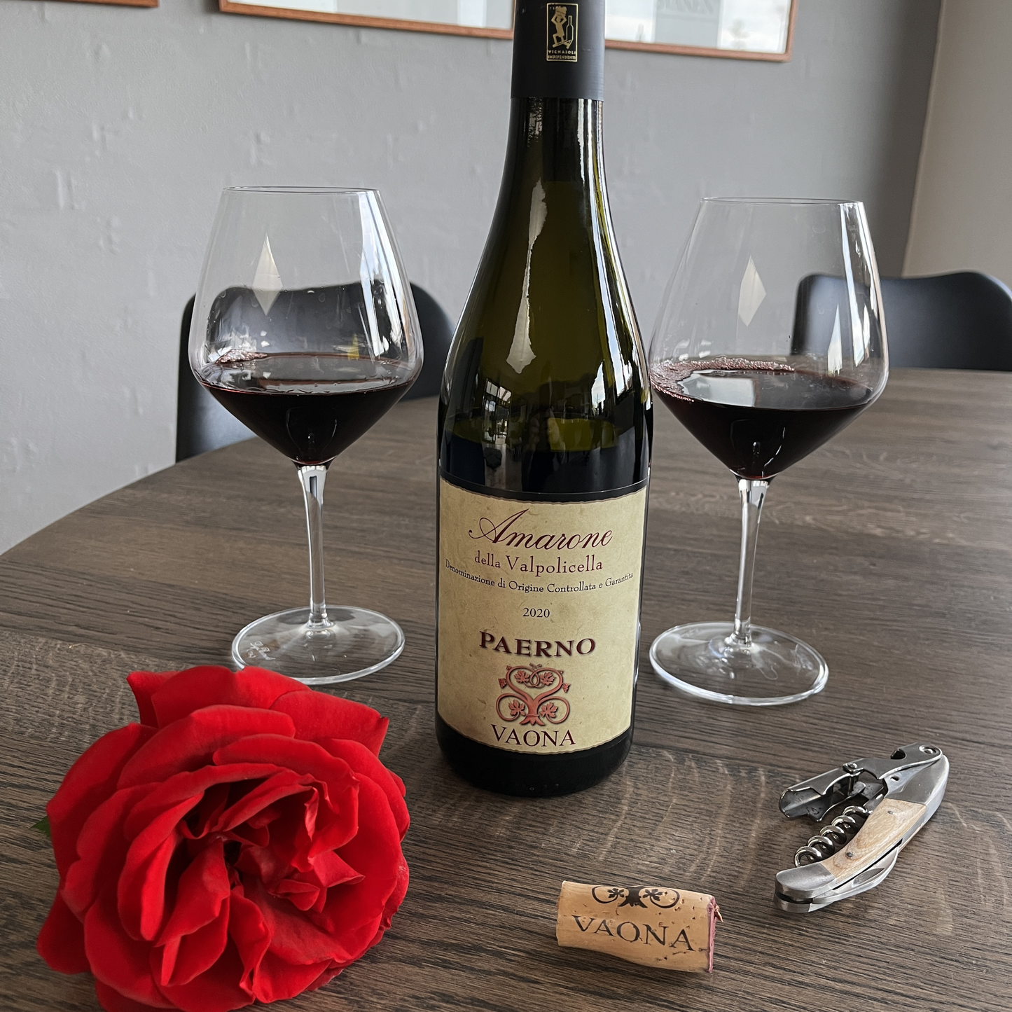 A rustic wooden table setup featuring a fresh red rose, a Vaona cork, and a wine opener in the foreground, with two glasses of red wine and the Vaona Amarone bottle standing slightly behind them.