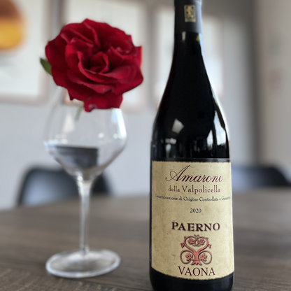 A bottle of Vaona Amarone Classico stands elegantly on a rustic table, with a blurred empty wine glass and a soft-focus red rose adding atmosphere to the background.