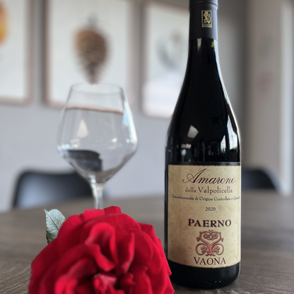 A close-up shot of a fresh red rose resting beside a bottle of Vaona Amarone Classico wine on a wooden table, featuring a subtly blurred empty wine glass behind it.