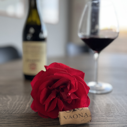 A detailed macro highlight of the authentic Vaona branded cork sitting in front of a red rose, with a glass full of red wine and the bottle visible in the warm, blurred background.